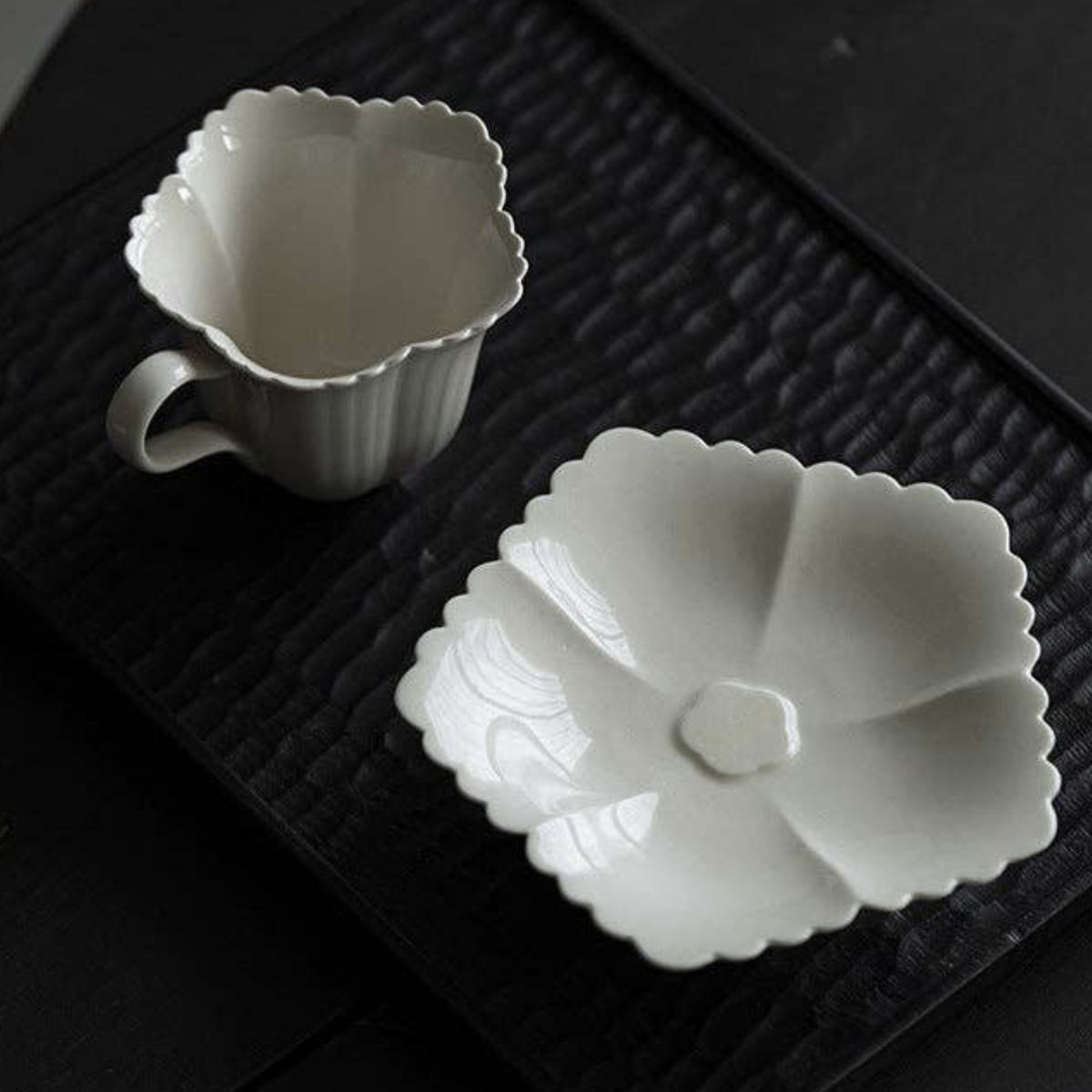 A white ceramic cup and matching flower-shaped saucer with scalloped edges, photographed on a dark textured tray against a dark background.