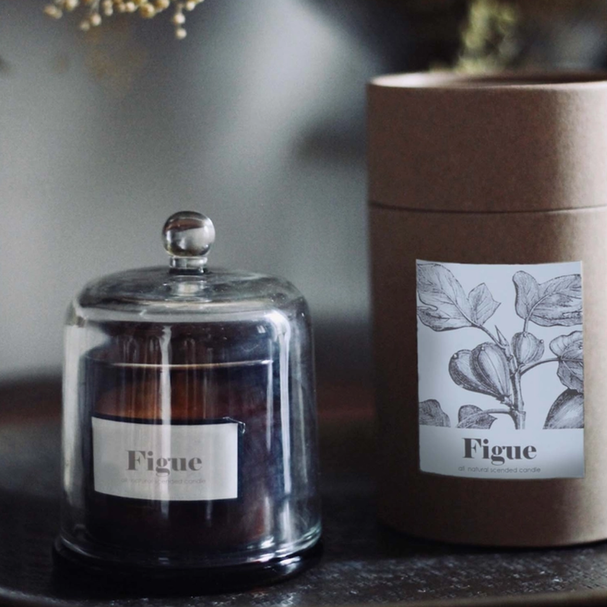 A dark amber glass jar candle under a clear glass dome with a silver knob, shown next to its brown kraft paper cylindrical packaging. Both pieces feature a white label with a botanical leaf illustration and the brand name 'Figue'.