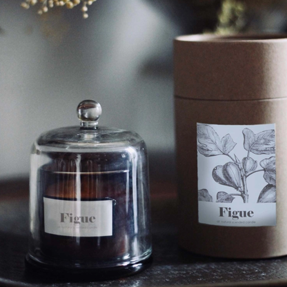 A dark amber glass jar candle under a clear glass dome with a silver knob, shown next to its brown kraft paper cylindrical packaging. Both pieces feature a white label with a botanical leaf illustration and the brand name 'Figue'.