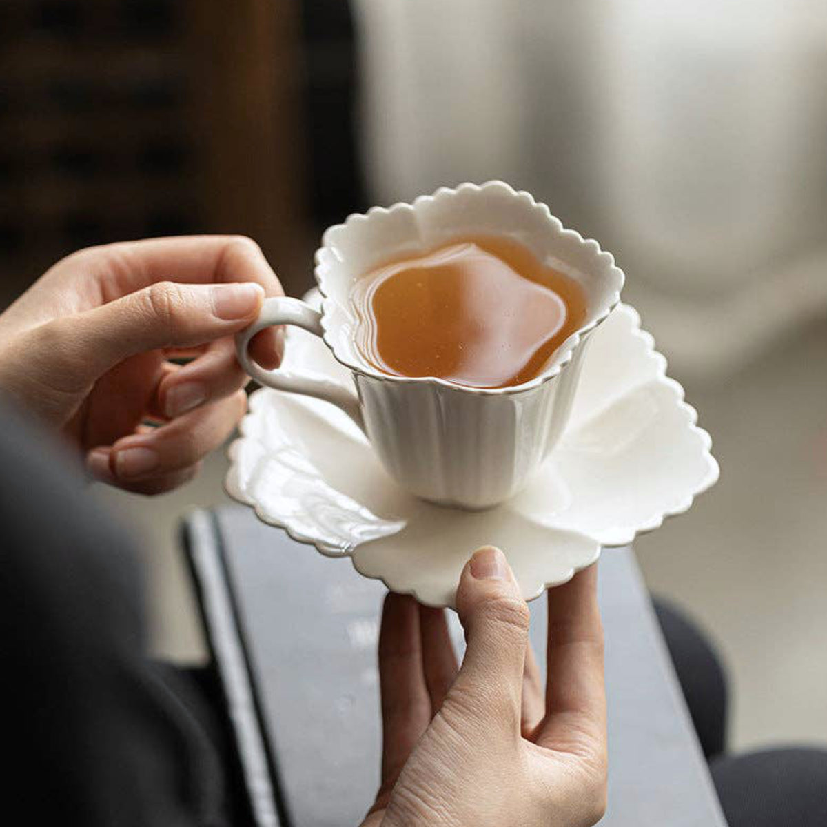 A person holds a white ceramic teacup and matching saucer with a scalloped edge, filled with amber tea, in a softly blurred indoor setting.