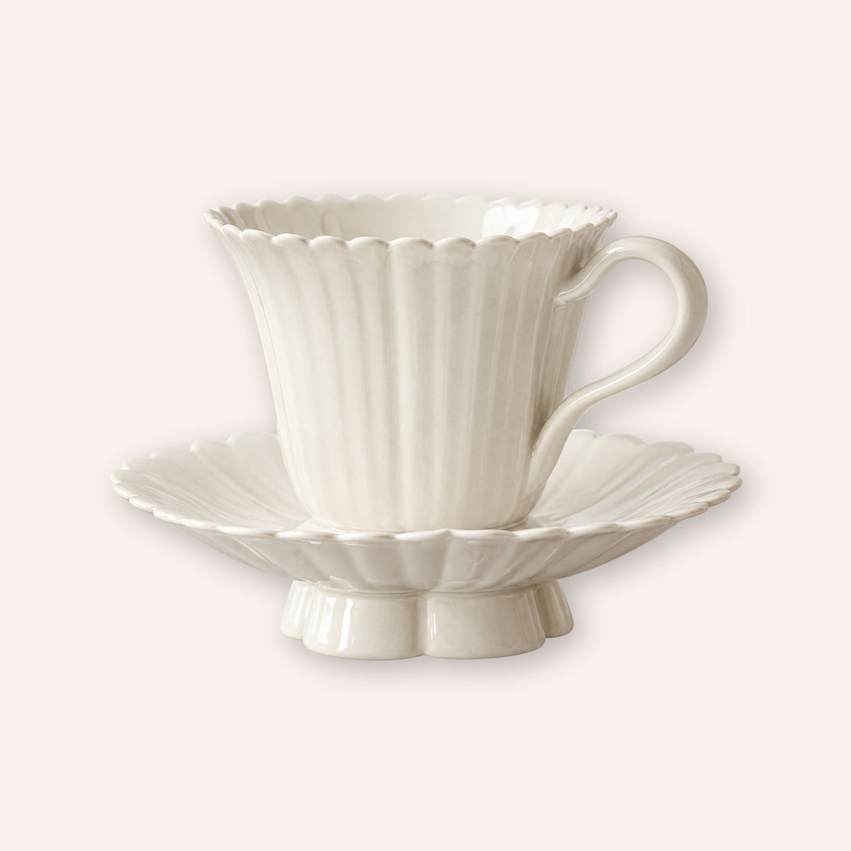A white ceramic teacup and saucer set with a fluted, scalloped edge design and a ribbed texture, set against a solid pale pink background.