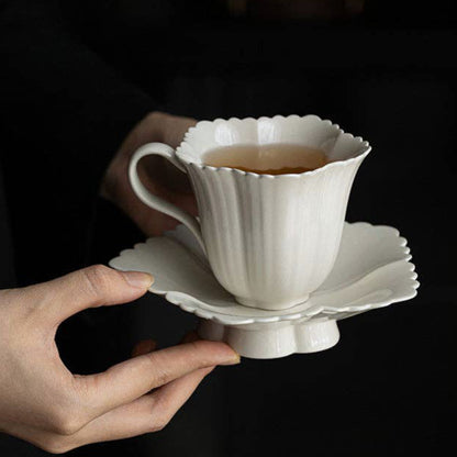 A white ceramic teacup and saucer set with a fluted, scalloped edge, held by hands against a dark background. The cup contains tea.