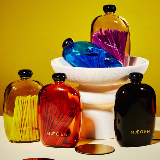 Five glossy glass match bottles with black caps from MÄGEN are arranged on a white surface against a mustard yellow background. The bottles come in amber, translucent red, and black, each filled with matching colored matchsticks.