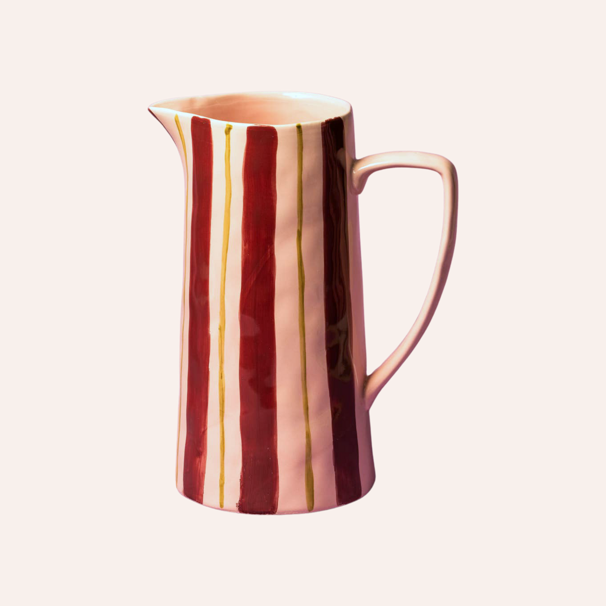 A ceramic pitcher with a glossy finish, featuring vertical stripes in deep burgundy red, soft pink, and thin gold lines, set against a solid pale cream background.