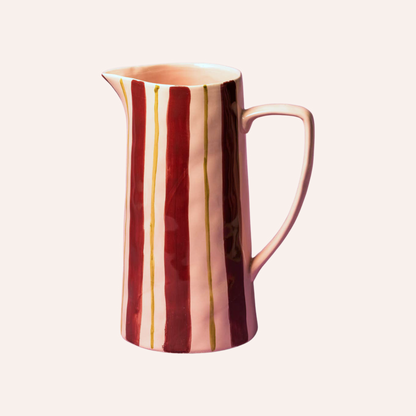 A ceramic pitcher with a glossy finish, featuring vertical stripes in deep burgundy red, soft pink, and thin gold lines, set against a solid pale cream background.