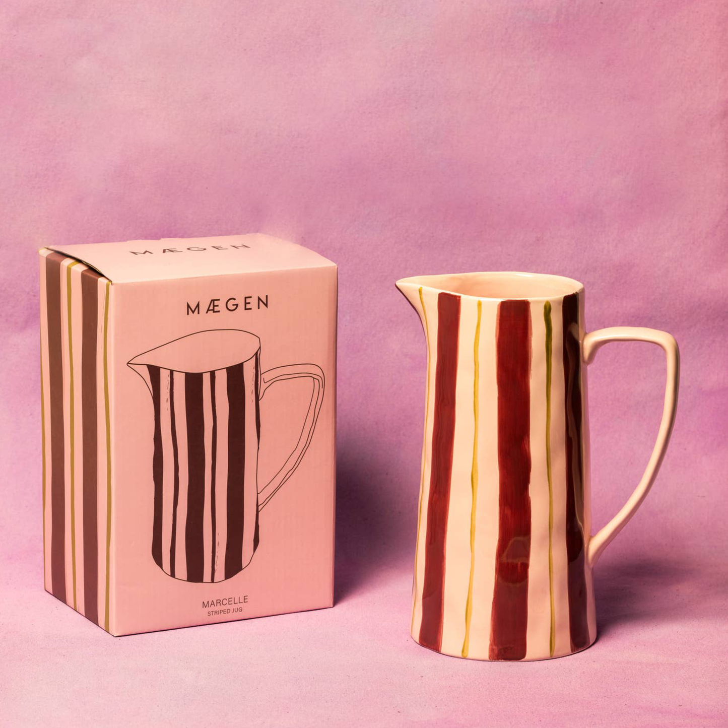 A striped ceramic pitcher with vertical wavy bands of cream, brown, and deep red, shown next to its pink and brown branded packaging box on a pink background.