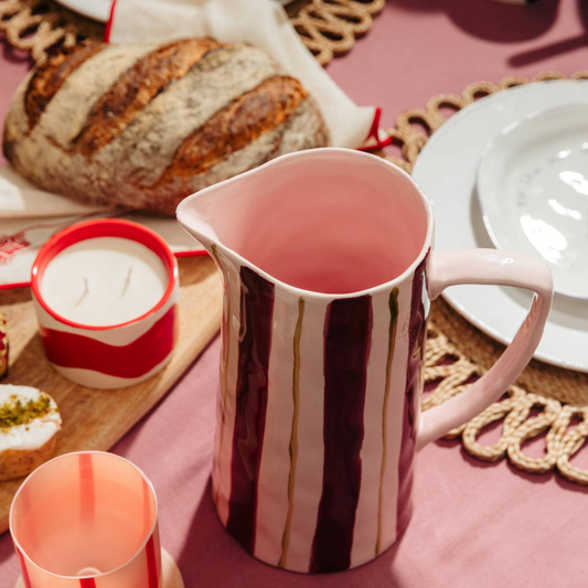 A pink ceramic pitcher with vertical dark purple and white stripes, styled on a pink tablecloth with bread, plates, and candles.