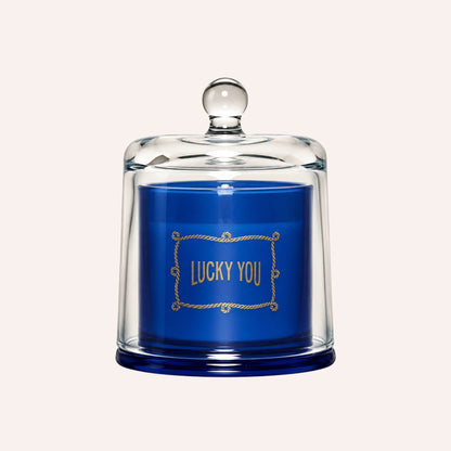 A blue candle under a clear glass cloche with a round knob lid, featuring gold foil text that reads 'LUCKY YOU' on the front.