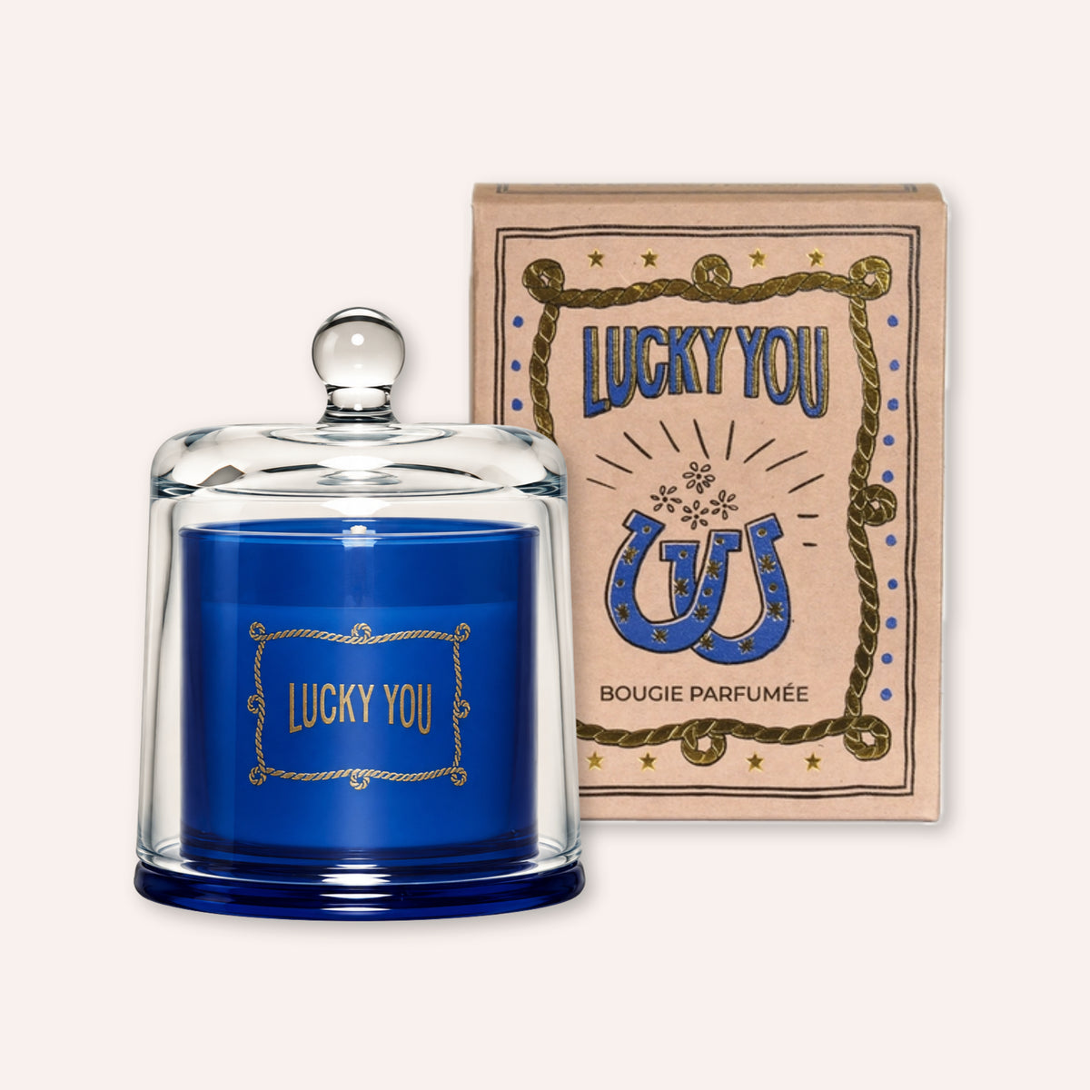 A blue scented candle in a clear glass dome with a glass knob lid, displayed next to its beige packaging box featuring a blue horseshoe and the text 'LUCKY YOU'.