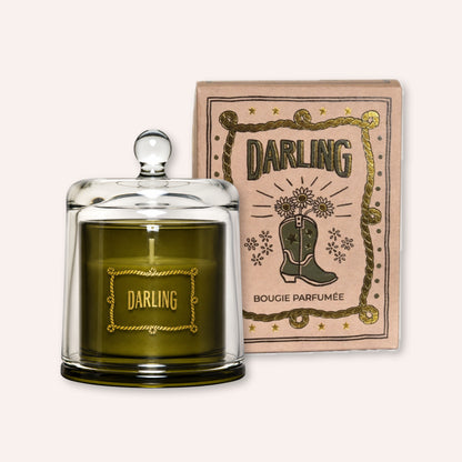 A scented candle in a clear glass jar with a glass lid sits in front of its illustrated kraft paper box. The candle wax is olive green, and the label reads 'DARLING'.