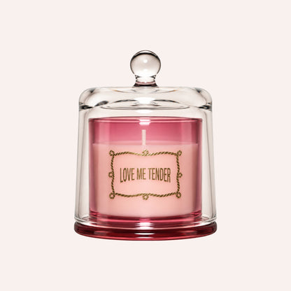 A pink candle inside a clear glass jar with a matching glass lid, featuring a gold decorative border and the text 'LOVE ME TENDER' on the front, set against a solid off-white background.