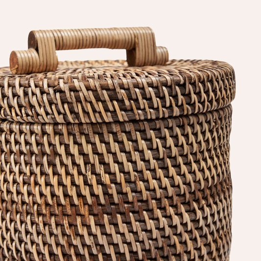 Close-up of a woven rattan or wicker storage basket with a matching lid and a curved wooden handle, shown against a plain light background.