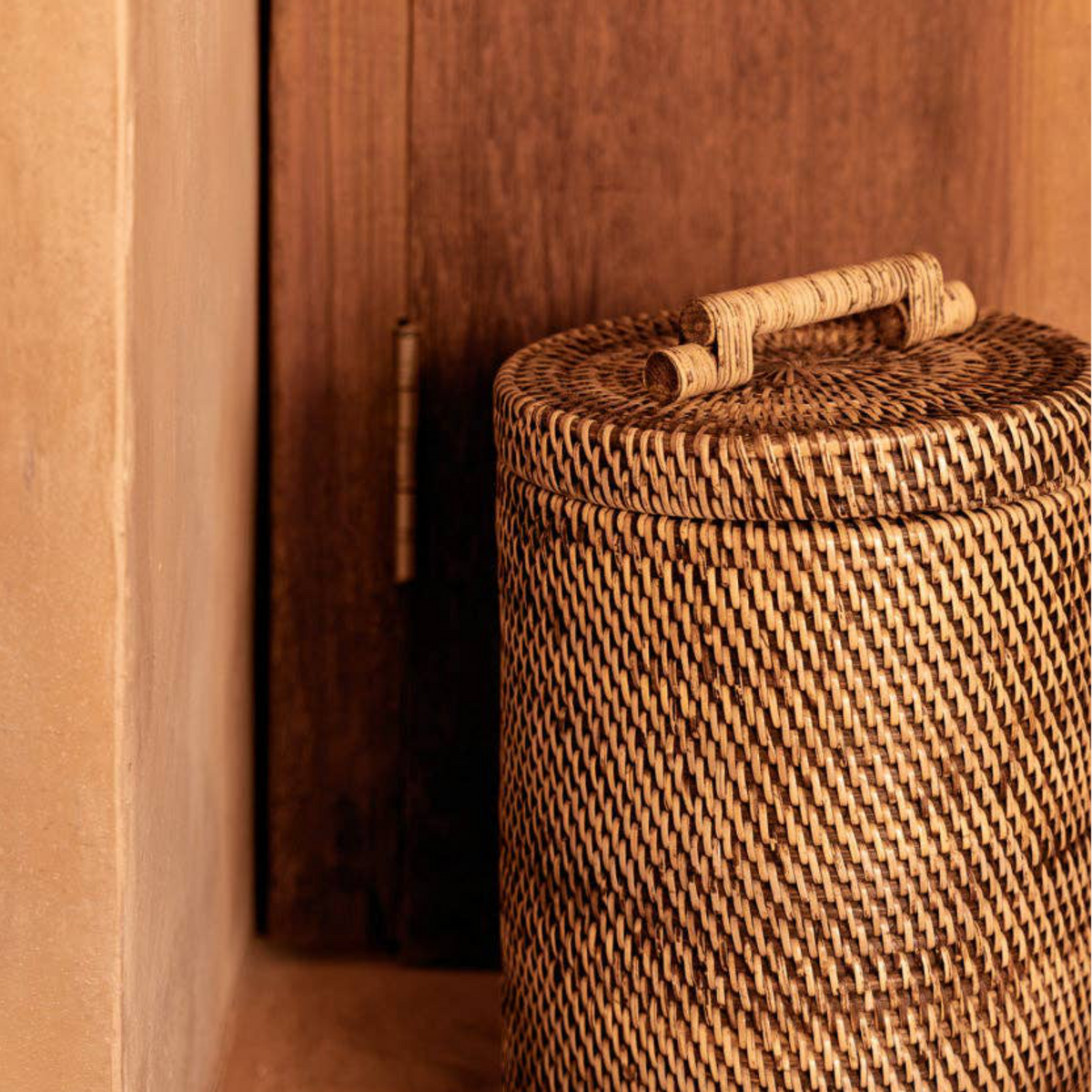 A close-up of a woven natural-fiber storage basket with a lid and integrated handle, placed beside a dark wooden door in a warm indoor setting.