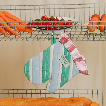 A striped fabric oven mitt hangs from a metal wire shelf rack in a kitchen setting, surrounded by carrots, tomatoes, eggs, and bread. The mitt features vertical stripes in light blue, mint green, and white on one side, and pink and white stripes on the other, with a small sewn-on label.
