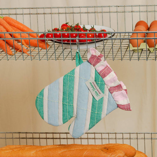 A striped fabric oven mitt hangs from a metal wire shelf rack in a kitchen setting, surrounded by carrots, tomatoes, eggs, and bread. The mitt features vertical stripes in light blue, mint green, and white on one side, and pink and white stripes on the other, with a small sewn-on label.