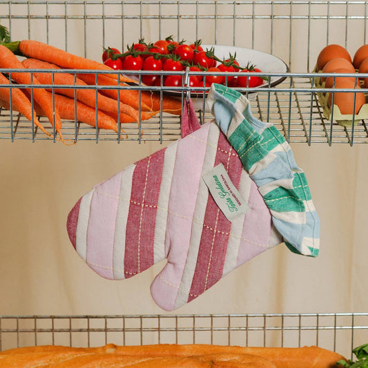 A striped fabric oven mitt with a ruffled cuff hangs from a metal wire rack in a kitchen setting, surrounded by carrots, tomatoes, and eggs.